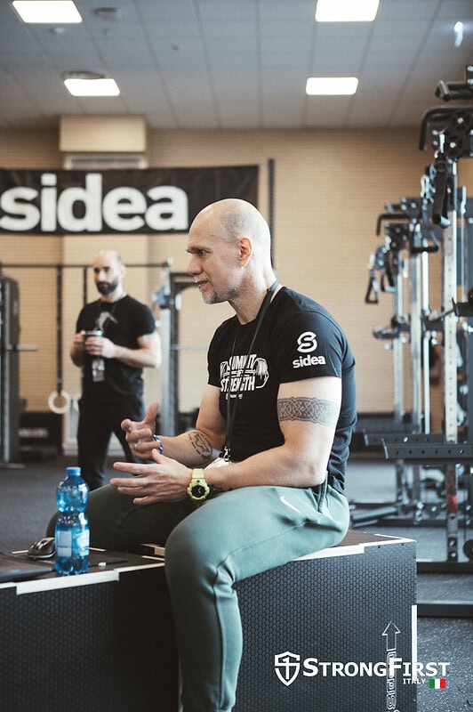 Allenamento in palestra con attrezzature professionali presso Sidea, azienda leader in attrezzatura fitness. Uomo seduto su panca, pronto a iniziare l'allenamento, con altri praticanti e attrezzature.