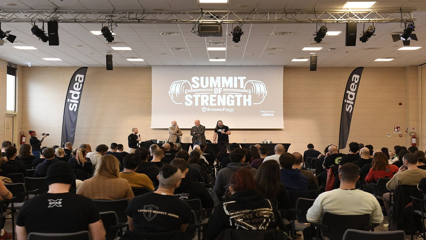 Gruppentraining in einem Fitnesscenter mit großem Publikum, Bühne mit Rednern und Leinwand mit dem Logo von "Summit of Strength", professioneller und motivierender Rahmen für F-Geräte-Fans.