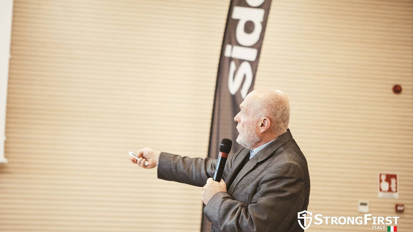 Uomo anziano con barba che parla durante una presentazione, tenendo un microfono e indicando uno schermo, in un evento di fitness professionale.