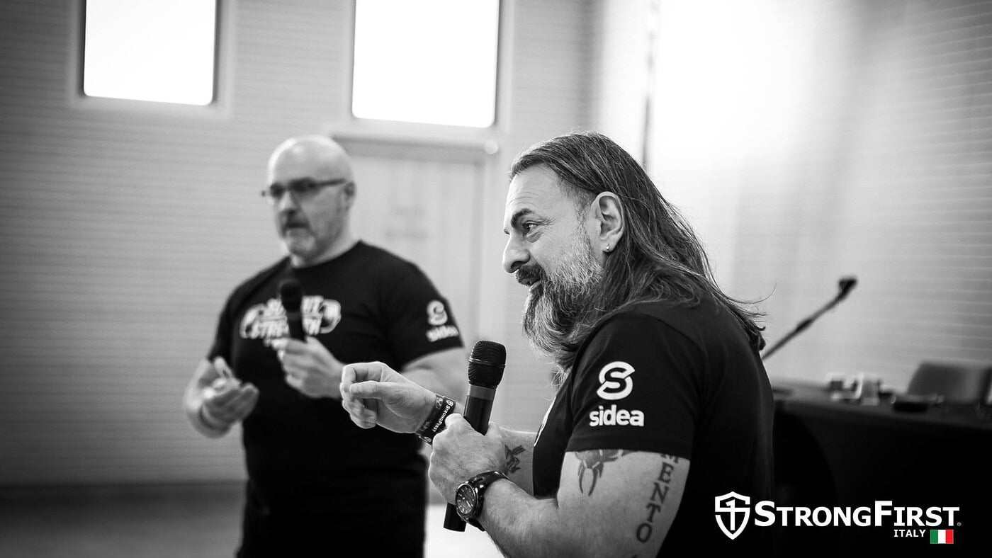 Due istruttori di fitness professionale durante un workshop, uno con microfono che spiega tecniche di allenamento e ipertrofia muscolare, in un ambiente di palestra.